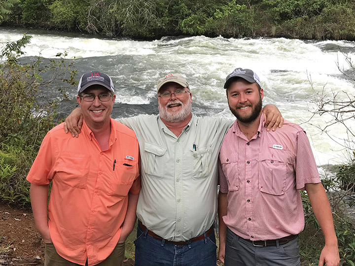 Dr. Gus Lorenz, Nick Bateman, and Dr. Ben Thrash pose for a photo. Dr. Lorenz stands between the two with his arms extended to the side and resting atop the shoulders of Mr. Bateman (left) and Dr. Thrash (right). They stand in the foreground with rushing river rapids just behind them. 